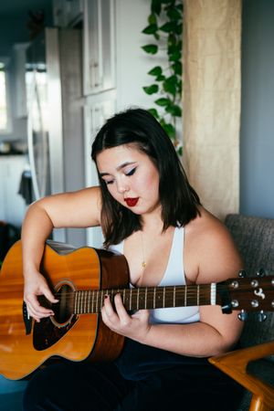 Young woman sitting on chair inside playing guitar