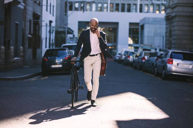 Handsome young man wearing suit talking on phone while walking with bicycle to work in the morning