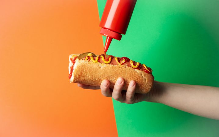 Person pouring ketchup on hot dog, on two tone background