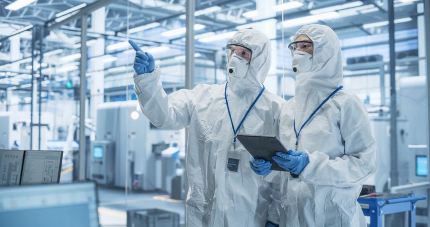 Bioengineers in Protective Coveralls, Masks and Eyewear Using Tablet Computer at a Chemical Manufacturing Complex. Laboratory Specialists Monitoring Conditions at a Modern Factory with Robotic Arms