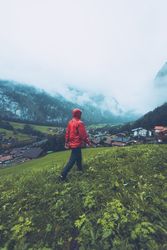 Back view of man in red jacket walking on green grass field near village in Switzerland bEJo10