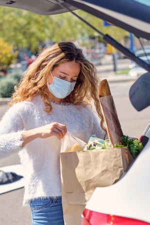 Woman in mask placing grocery bags in car trunk