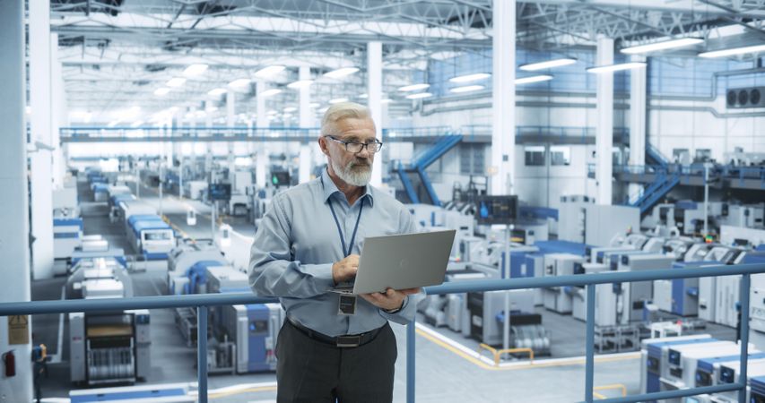 Engineer Standing on a Platform, Using Laptop Computer at an Electronics Factory. Machines are Undergoing Maintenance, Specialist Monitoring the Progress Through Online Software