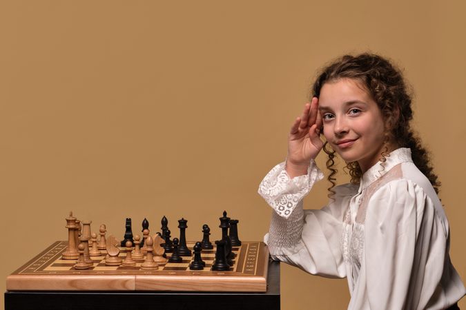 Girl smiling while raising hand in gesture near chessboard.