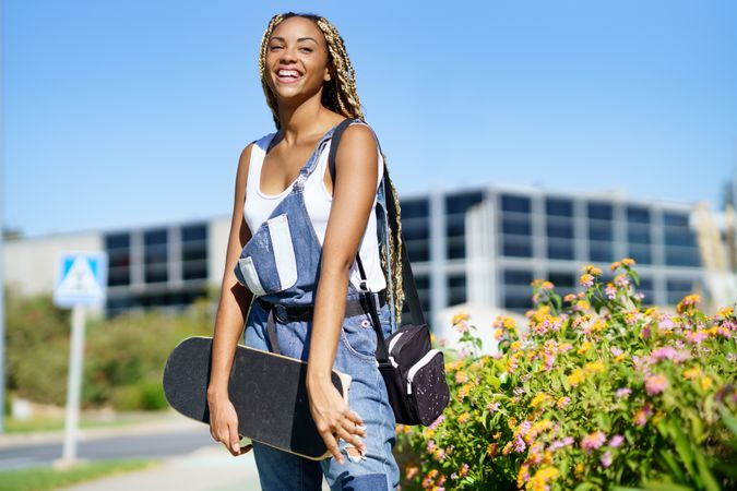 Smiling female in denim overalls holding skateboard on campus