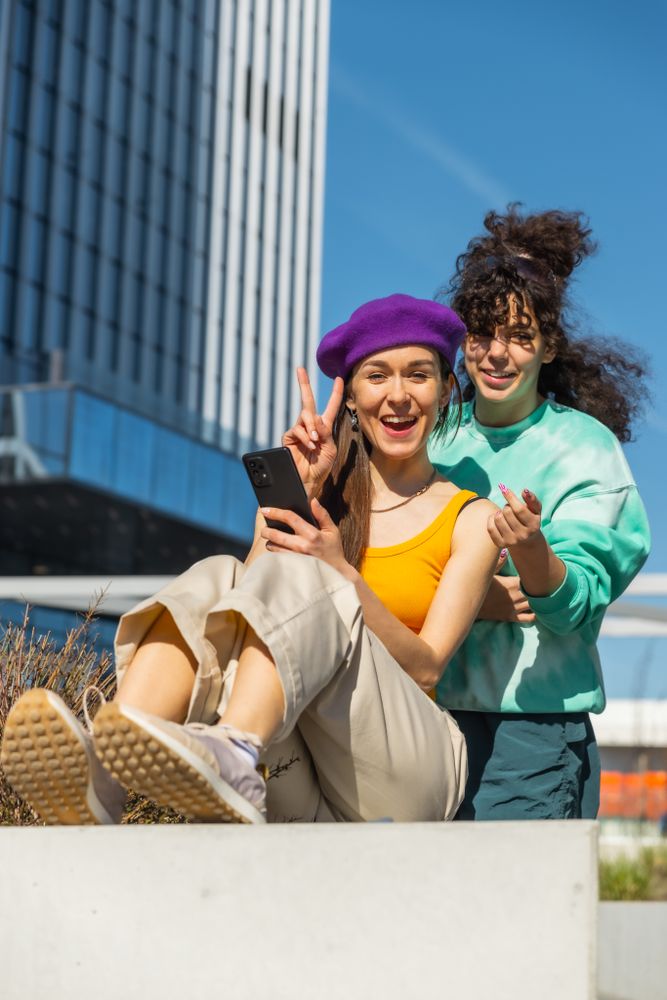 Vertical Screen: Two Young Women Share A Moment Of Laughter, Engaging With Smartphone Outdoors, Looking at Camera And Showing Peace Sign.