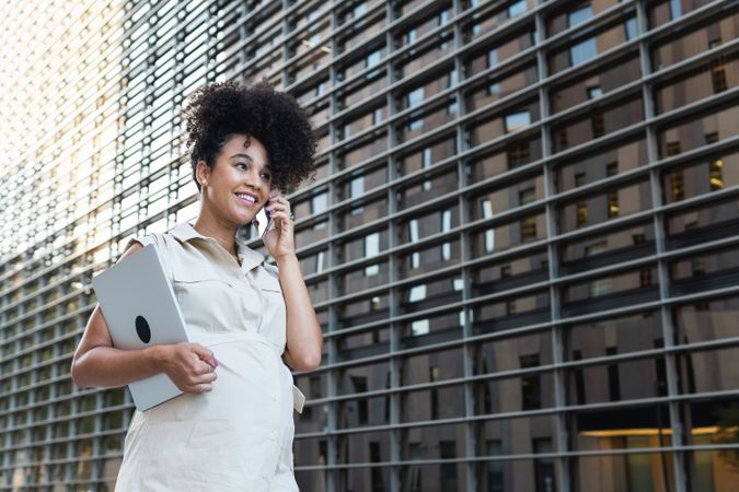 Smiling Black pregnant woman talking on smartphone and carrying a laptop