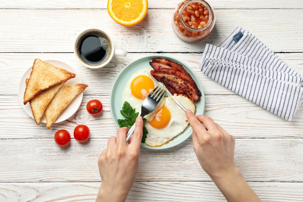 Top view of person’s hands cutting into an egg and bacon breakfast