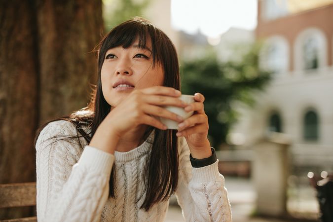 Woman sitting at outdoor table holding coffee and looking away