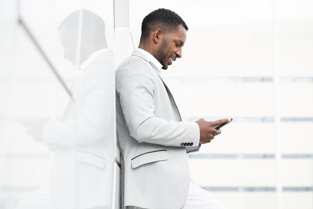 Elegant Black man browsing smartphone leaning on geometrical wall