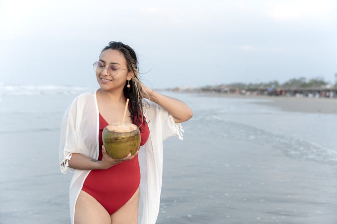 A young Hispanic woman is posing with a coconut on the sea edge at the beach