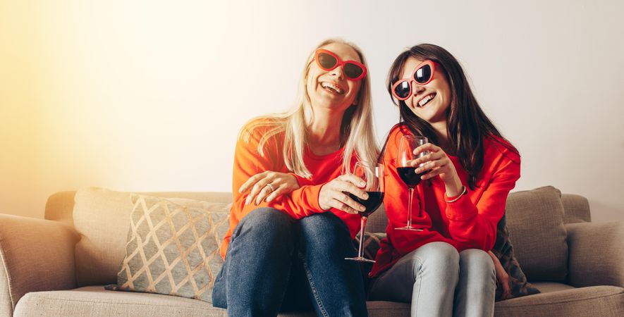 Older mother and adult daughter wearing sunglasses toasting with wine inside at home
