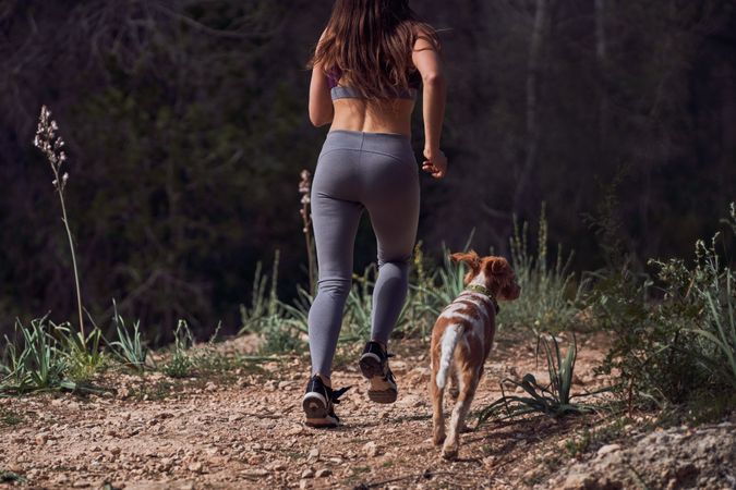 Crop fit sportswoman jogging with dog on narrow path in green forest
