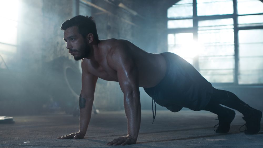 Muscular Shirtless Man Covered in Sweat Does Push-ups in a Deserted Factory Remodeled into Gym.