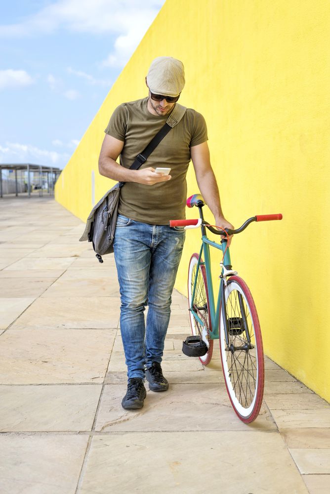 Man in hat and sunglasses walking along yellow wall and checking phone