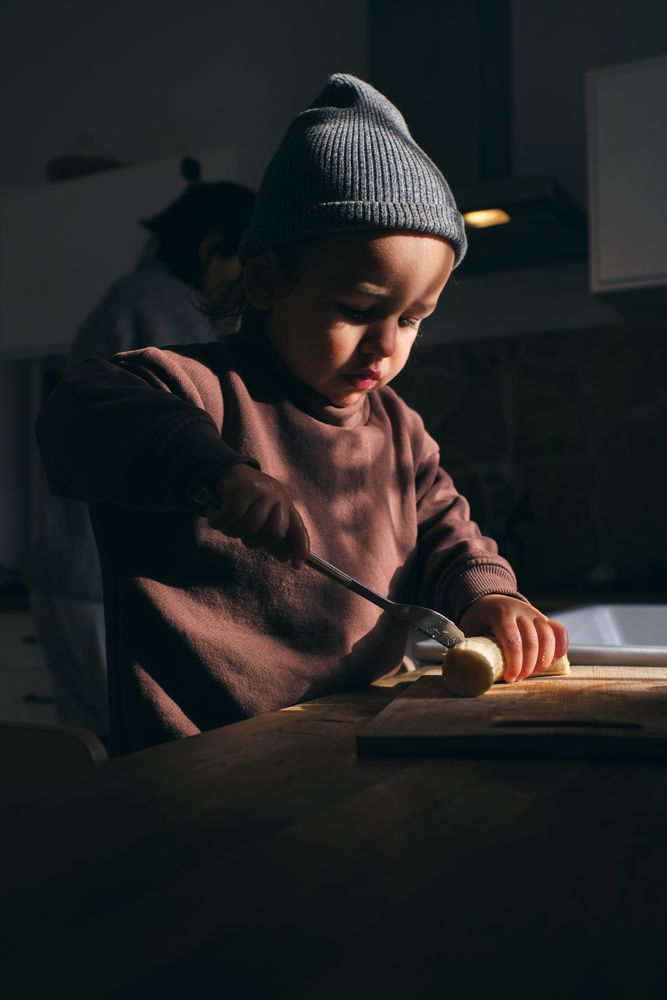 Serious toddler cutting fresh banana in kitchen