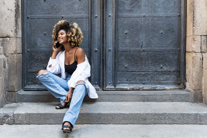 Delighted Black woman speaking on smartphone while sitting near building