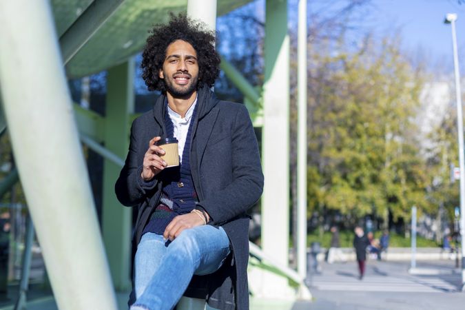 Front view of cheerful Black man in jacket with warm beverage balancing between poles outside