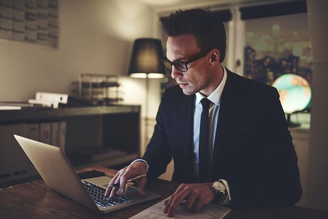 Man in suit and glasses working on his laptop and documents at night