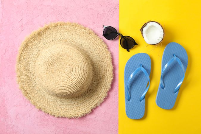 Straw hat, flip flops, sunglasses and coconut on two tone background, space for text and top view. Summer vacation backdrop