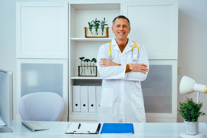 Mature doctor standing and smiling in a clinical setting with his arms crossed