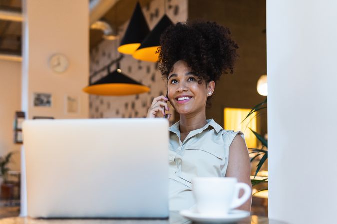Smiling Black woman talking on smartphone at table with laptop