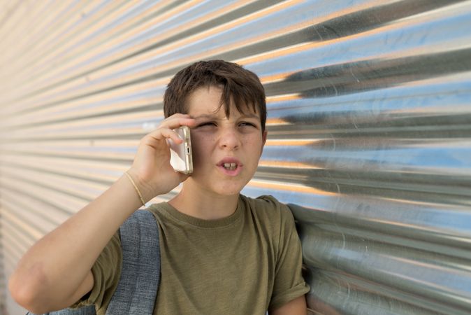 Boy having conversation on smartphone leaning on metal shutter