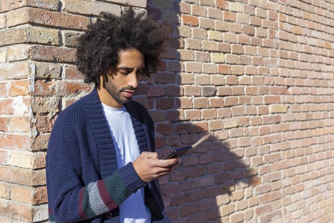 Man looking at his smartphone while leaning on a brick wall outdoors on sunny day