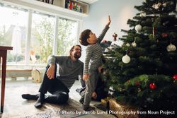 Small family together at home during Christmas with boy reaching up Christmas tree 5ry220