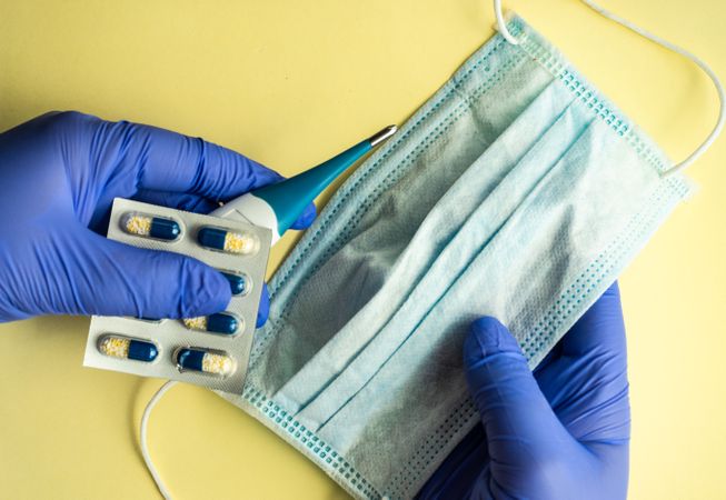 Top view of yellow table with hands wearing protective blue gloves holding facemask and pills