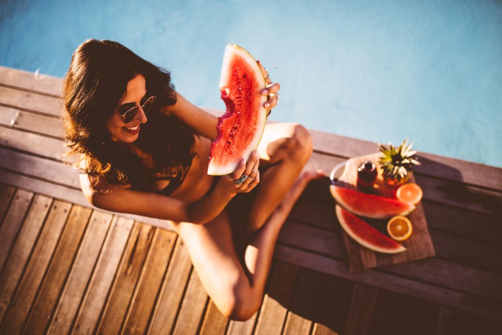 Smiling young woman sitting poolside with a fruit platter eating a slice of watermelon
