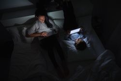 Woman and boy lying on bed and surfing gadgets at night