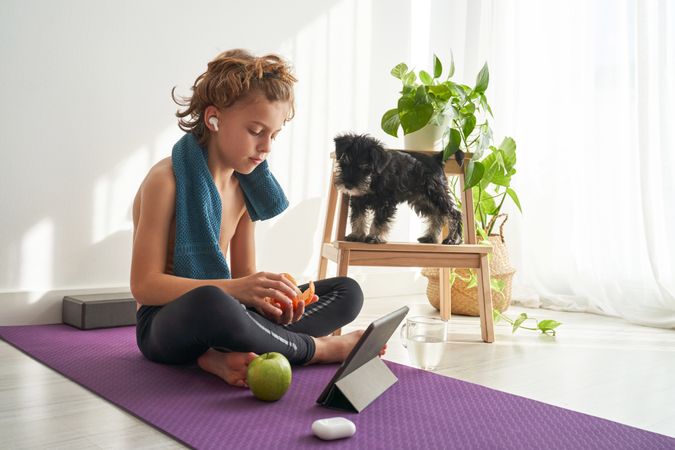Kid using tablet on mat and peeling orange near dog
