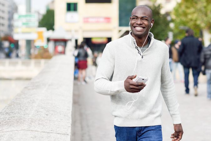 Male with shaved head wearing casual sweater walking with headphones and phone