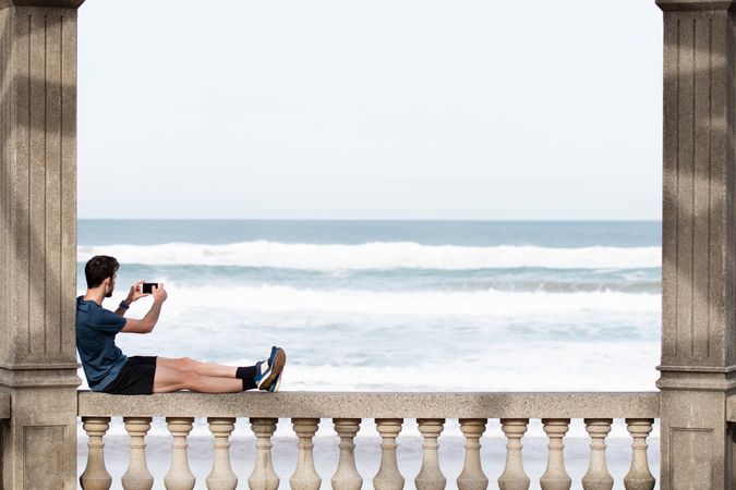 Man in sports clothing relaxing on fence and taking picture of sea on smartphone
