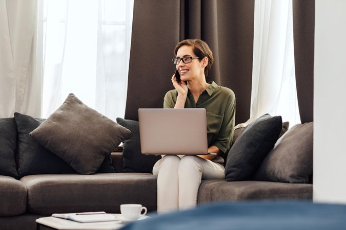 Smiling woman on sofa with pillows with cell phone and laptop