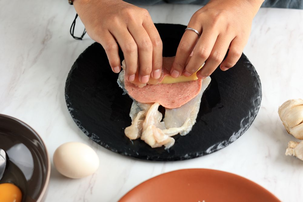 Person rolling salami slice with cheese and chicken breast