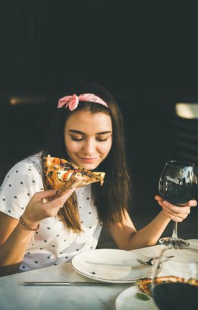 Young woman with red wine in hand, eating pizza outside in the sun, copy space