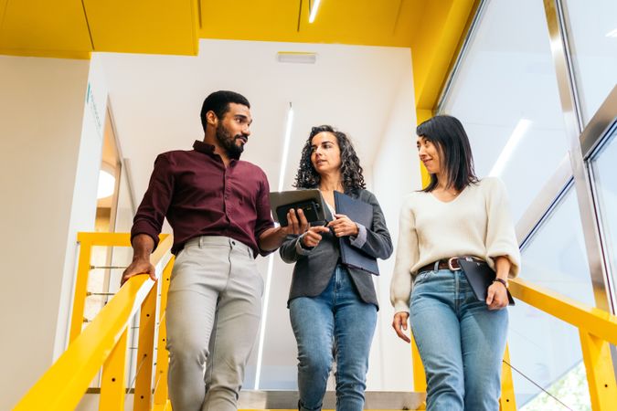 Multiracial coworkers happily talking about the company while walking down some yellow stairs in the office corridor