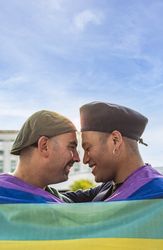 Gay couple under LGBT rainbow flag while laughing and looking at each other.