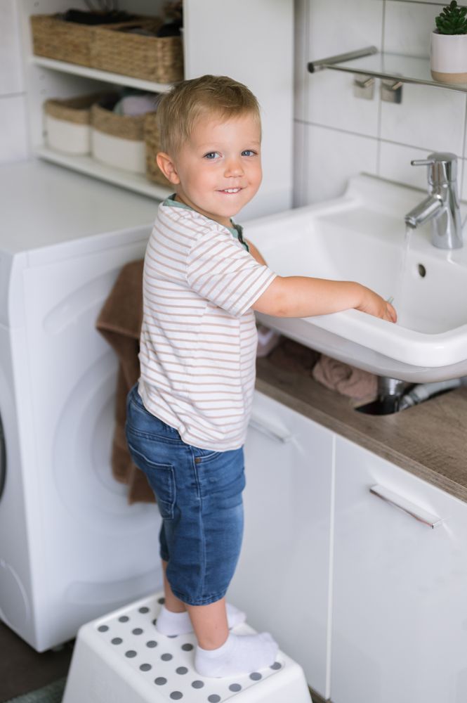 Cute child boy standing on stool near washing sink in daylight