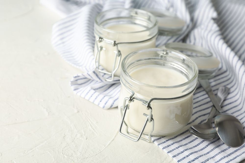 Glass jars with sour cream yogurt, spoons and towel on cement background