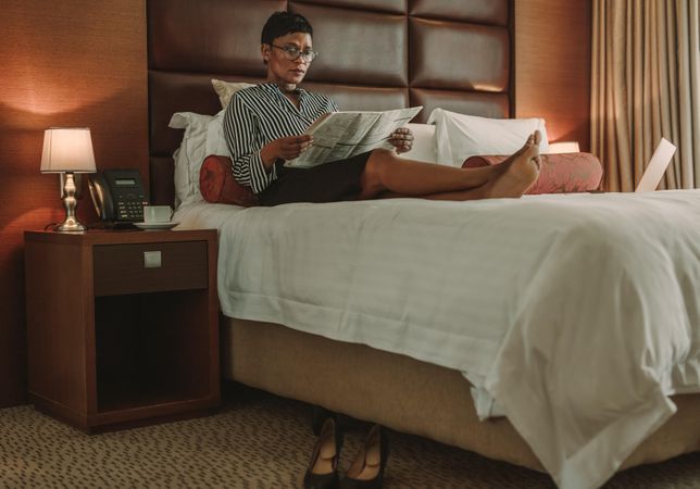 Businesswoman relaxing on bed reading newspaper in modern hotel room
