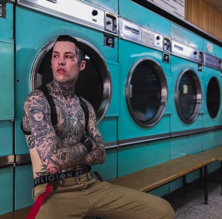 Young tattooed shirtless man sitting with crossed arms in laundromat