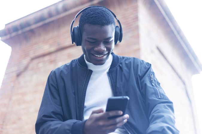 Smiling Black man outside, wearing headphones and checking his phone