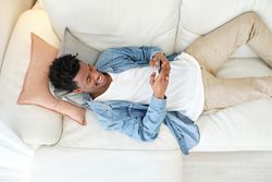 Happy Black man resting on sofa and using smartphone