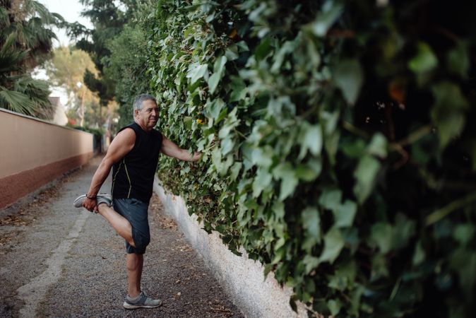 Mature man stretching leg holding botanical wall in street