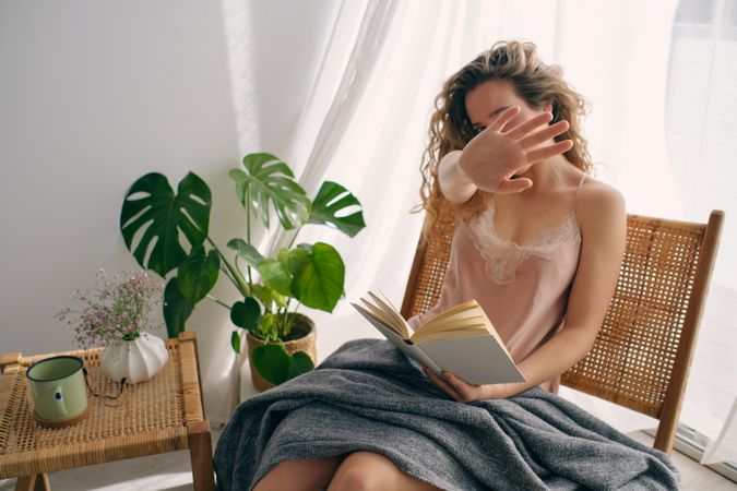 Young woman reading book on wicker chair and hiding behind hand