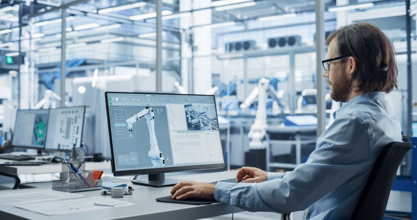 Portrait of a Young Factory Manager Working on Desktop Computer in Industrial Manufacture with Autonomous Assembly Robots. Production Line Making Heavy Machinery Parts in the Background