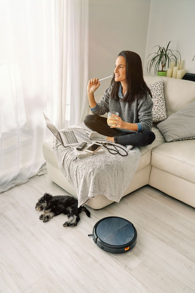 Smiling female sitting on sofa near sleeping dog and robotic vac